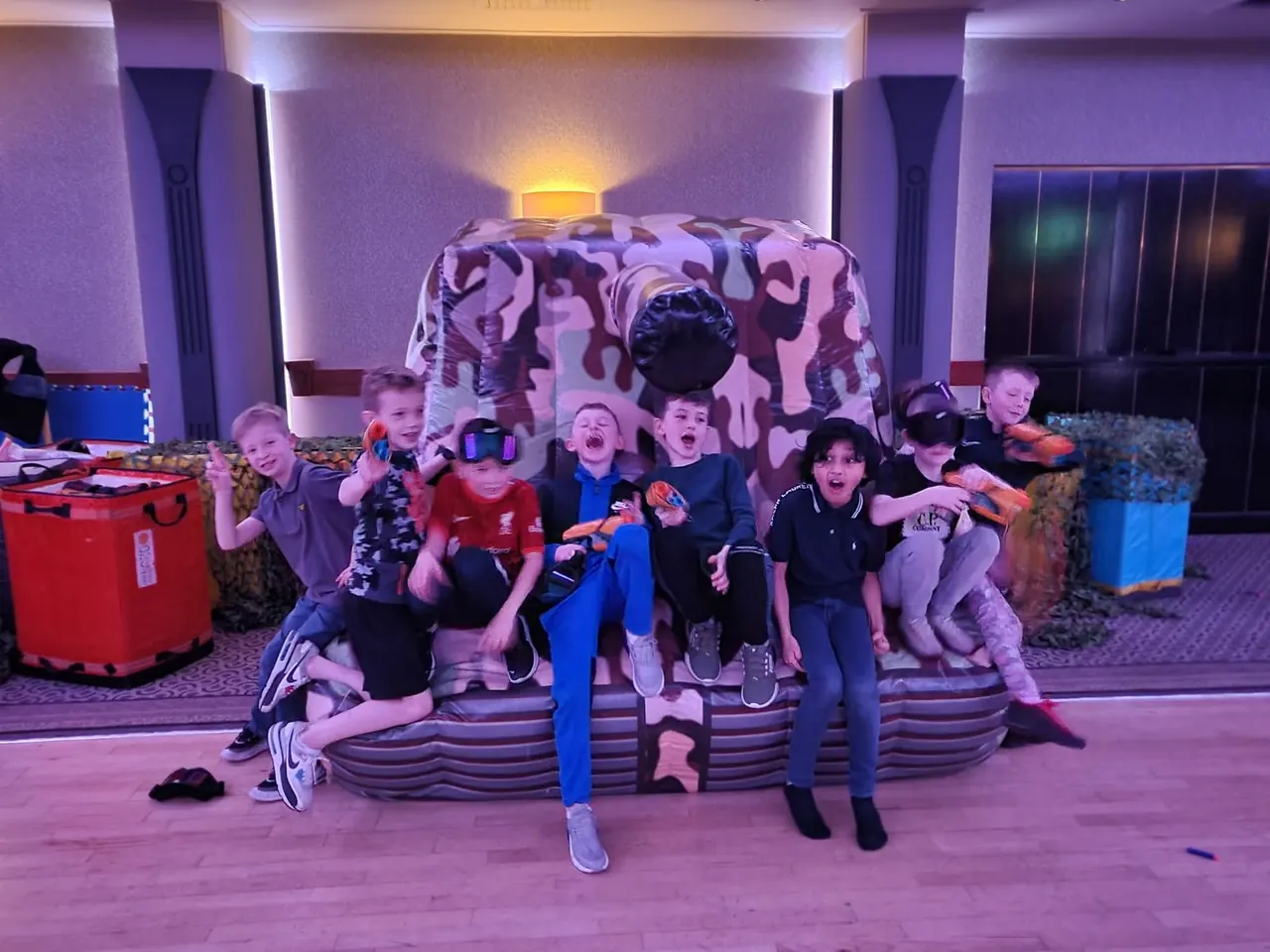Boys posing with blasters around the inflatable tank in a hotel function suite lit with party lighting
