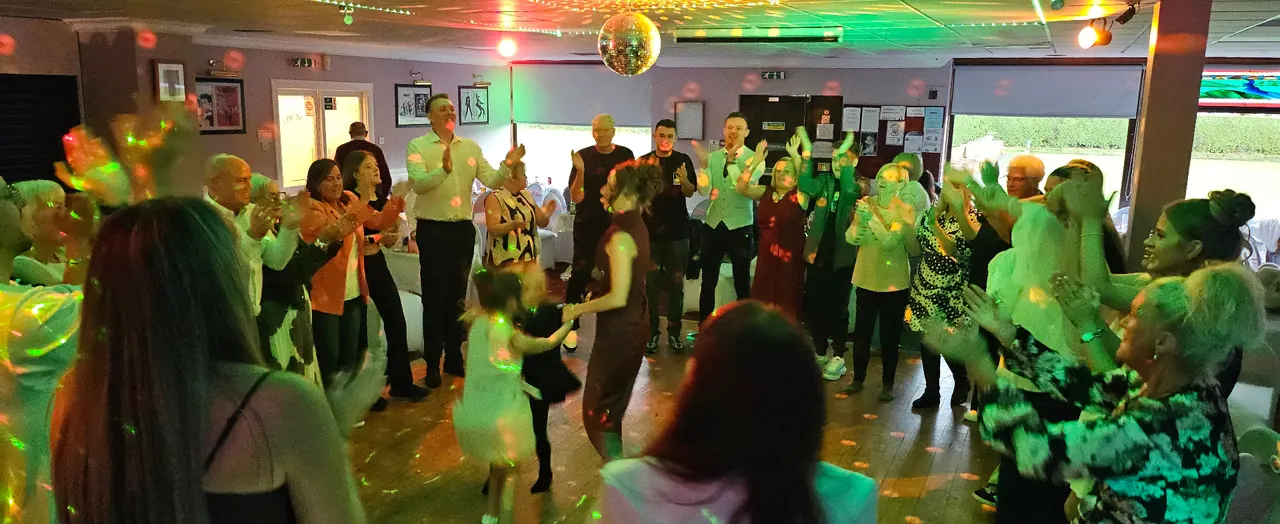 A packed dance floor of children and adults clapping along under party lighting at a family celebration