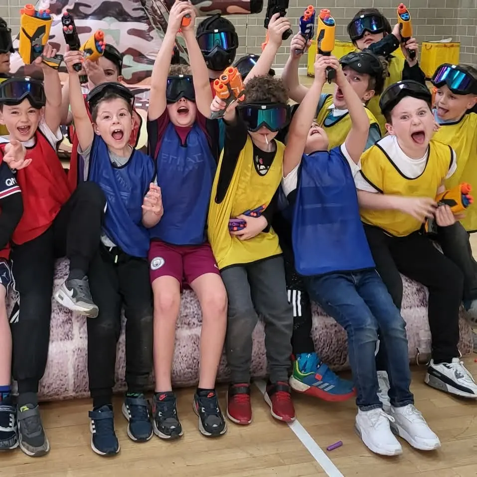 A team of children in coloured bibs and safety goggles cheering with foam blasters held in the air