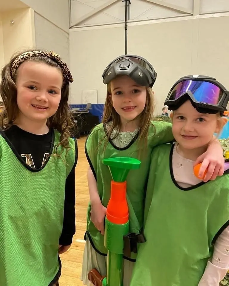 Three girls in green team bibs with goggles on their heads, holding a foam blaster in a hall