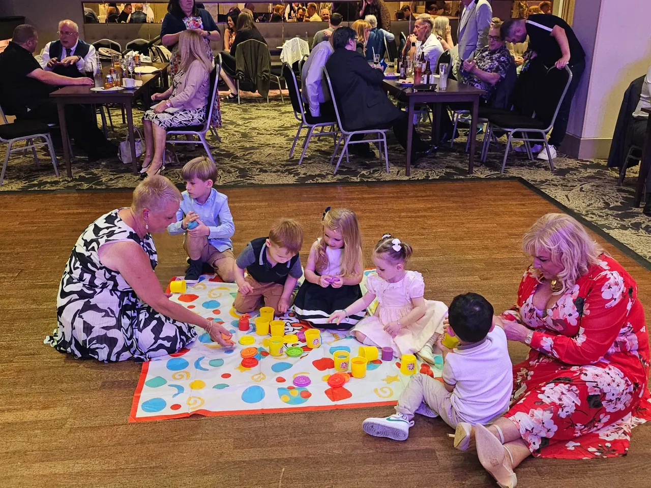 Toddlers and adults playing a floor game together while guests dine at a family celebration