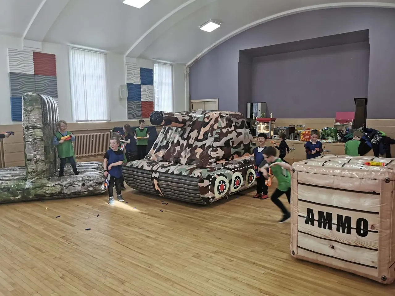 Children in team bibs running between an inflatable tank and an ammo crate during an indoor Nerf battle